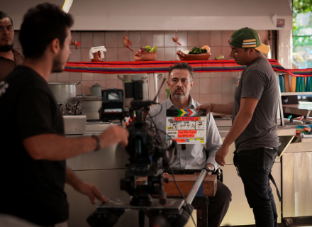 Fotografía del proceso de producción de un video. Hombre sentado en una cocina ve directamente a la cámara.